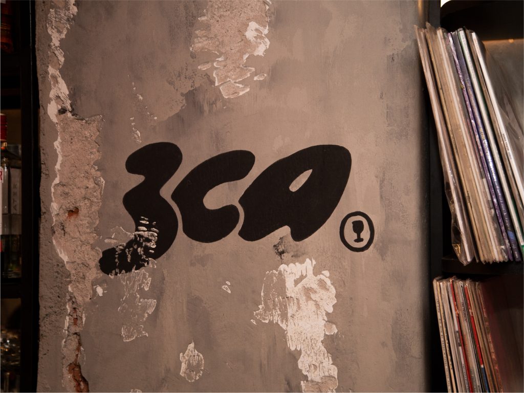 Detalle del logotipo de Tresca Vinilos y Copas pintado sobre una pared con revoque desgastado, junto a una estantería llena de vinilos.