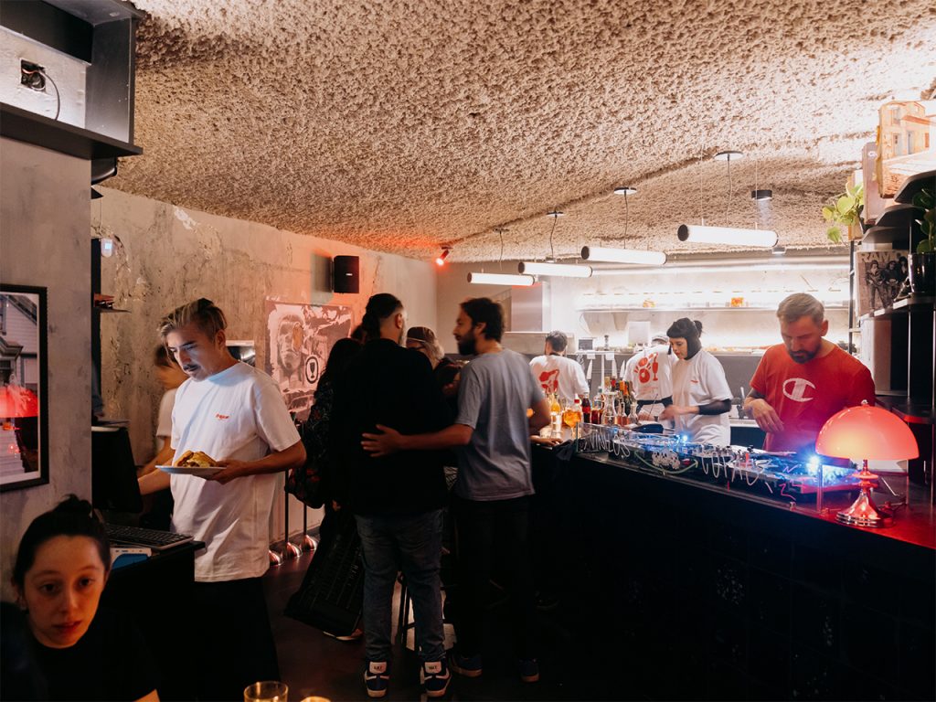 Vista del interior del bar Tresca durante una noche de servicio. Se observa la barra en primer plano con una lámpara roja encendida y una bandeja de DJ. Al fondo, el equipo trabaja en la cocina mientras varias personas conversan y disfrutan en un ambiente cálido y texturado.