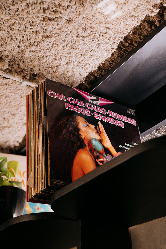 Primer plano de un vinilo de música latina con portada retro, en una estantería negra bajo un techo texturado. El título del disco destaca en letras fucsias: "Cha Cha Chas · Rumbas · Pasos · Sambas".